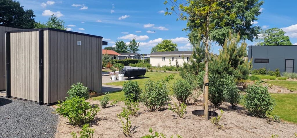 Tuin met groene grasvelden, plantenbedden en houten verblijven bij EuroParcs Bad Hoophuizen onder een blauwe lucht.