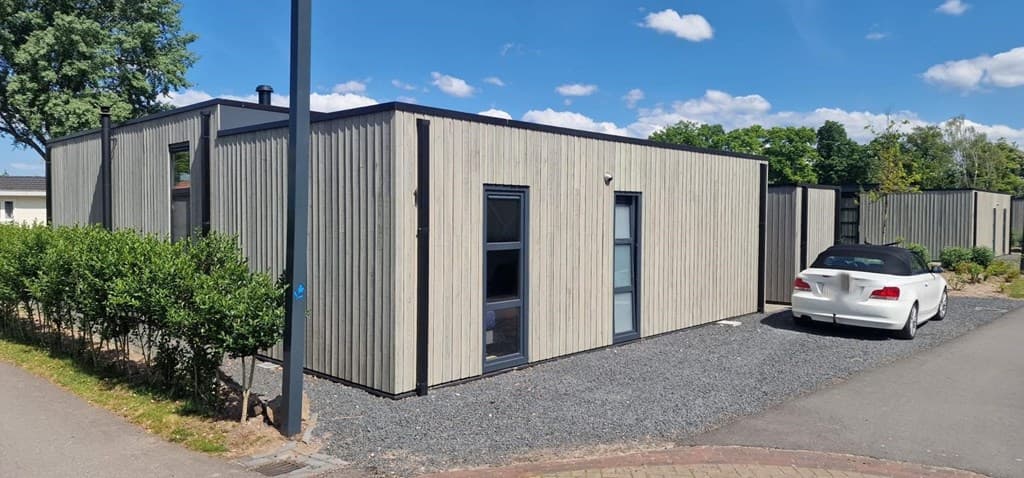 Moderne vakantiewoning met een houten gevel, omringd door groen en een grintpad met een witte auto voor de deur