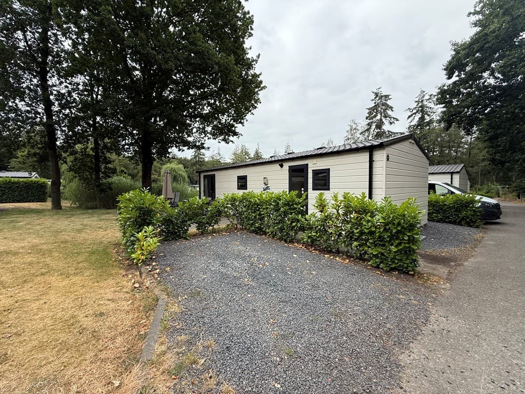 Zicht op een bungalow met een omheining van hagen, omringd door bomen en een grindpad met een geparkeerde auto.