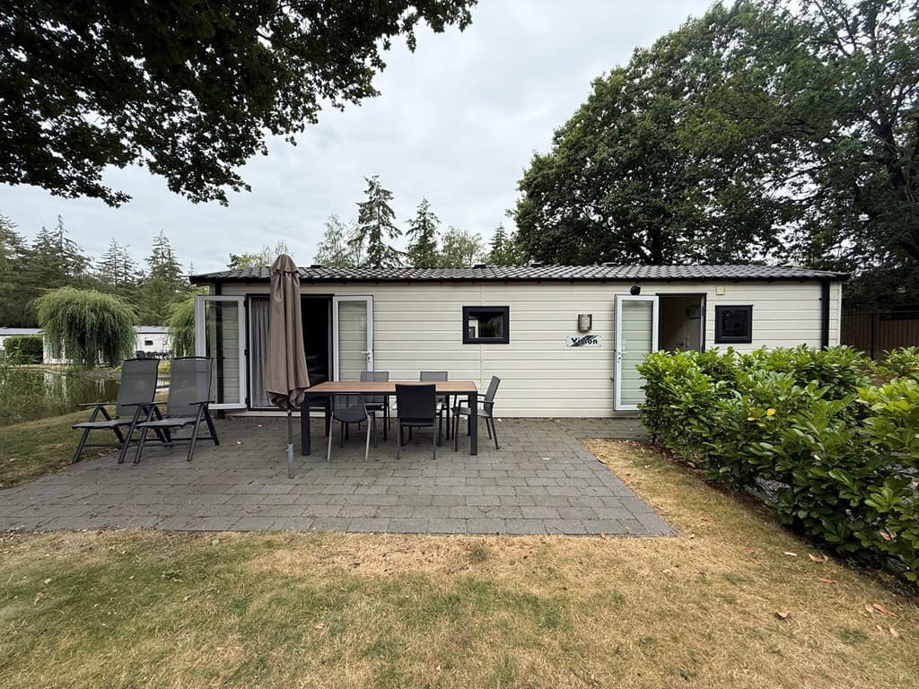 Houten vakantiewoning met terras en tuinmeubilair, groene omgeving met bomen en grasveld.