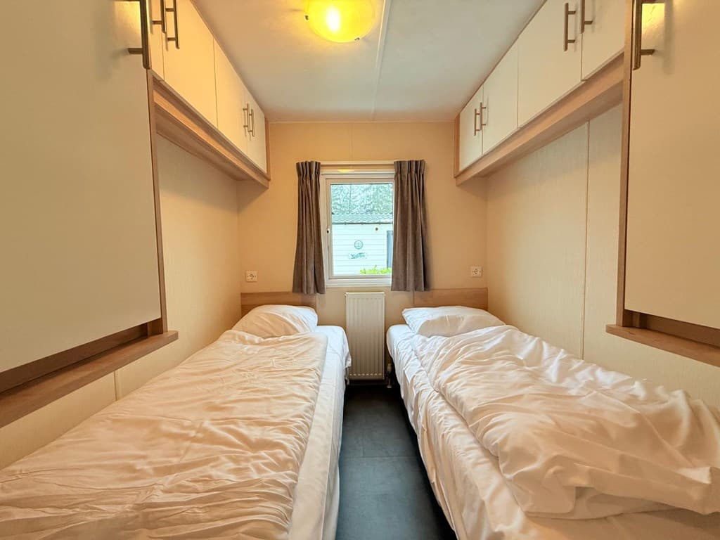 Slaapkamer met twee eenpersoonsbedden en opbergruimte boven, raam met vitrage voor natuurlijk licht.