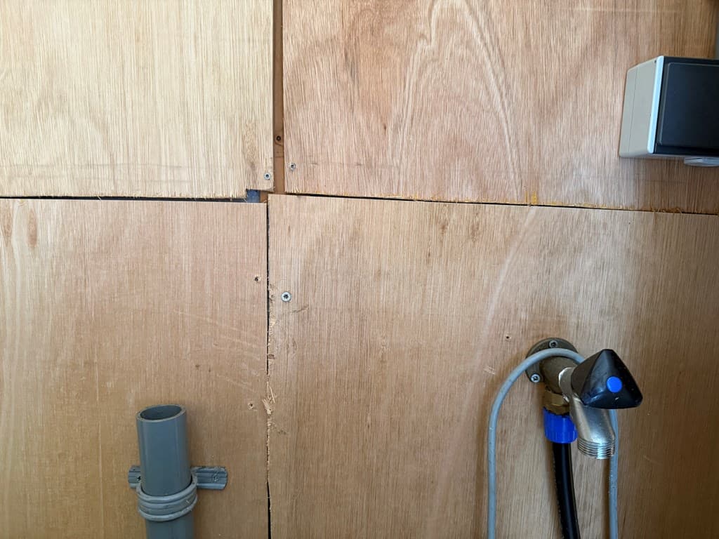 Houten wand met naden en een wateraansluiting, zichtbaar leidingen en een soort ventiel aan de rechterkant.
