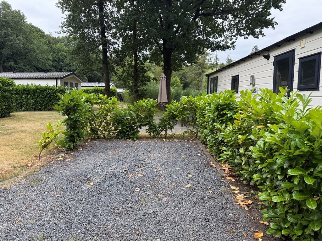 Entree van de woning met grindpad en groene haag, ruimte tussen de bebouwing en een parasol in de achtergrond.