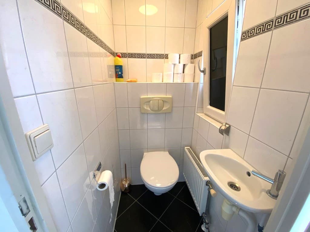 Klein toilet met wandtegels en een wastafel, voorzien van een raam en zwarte tegelvloer.