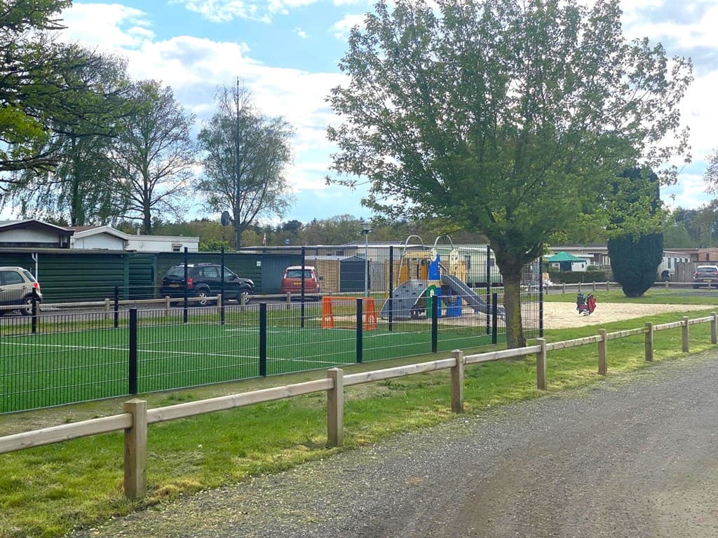 Speelplaats met speeltuin en zandbak, omgeven door groen en woningen, zicht op parkeerplaatsen en omheining.