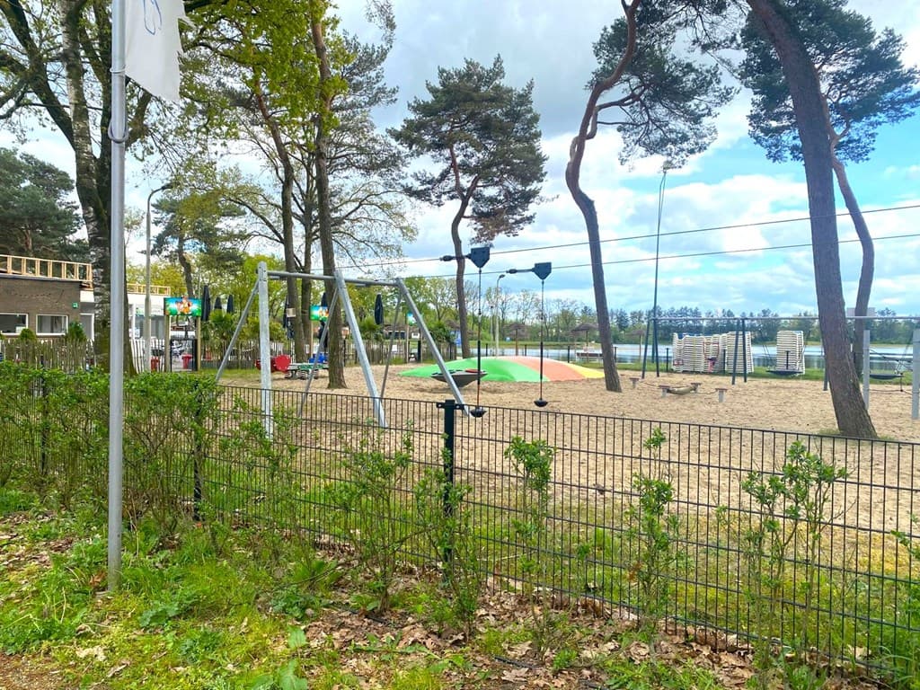 Speeltuin met schommels en een zandbak, omringd door bomen en een hek, met uitzicht op een waterpartij.