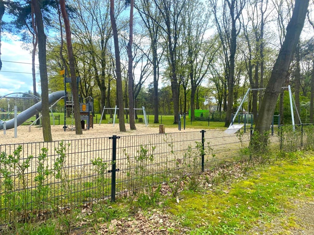 Speelplaats met zand, glijbaan en schommels, omringd door bomen en een hek aan de rand van een vakantiepark