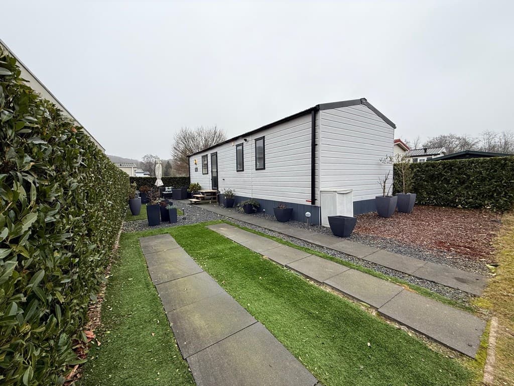 Woning met grijze buitenzijde, groene grasstrook en betegeld pad, omringd door hagen en plantenbakken.