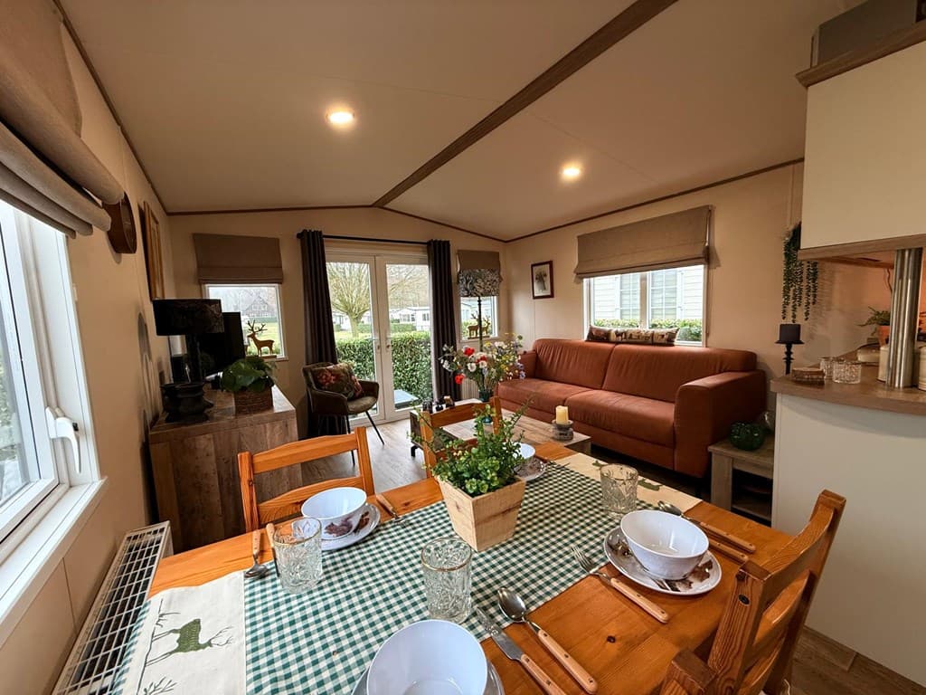 Eetkamer met houten tafel, glazen, servies en een bankenhoek; raampartij met uitzicht op de tuin.