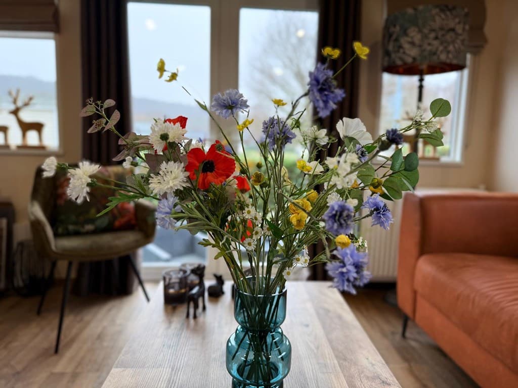 Vaas met een boeket diverse bloemen op een houten tafel in een woonkamer met grote ramen en een bank.