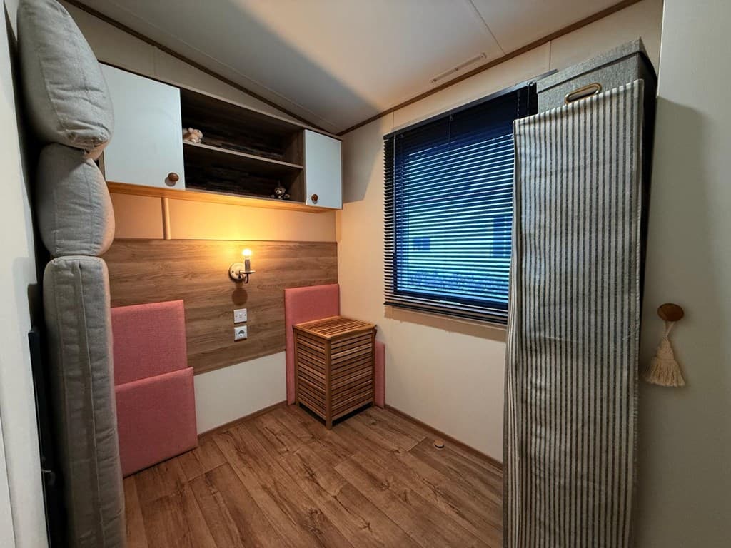 Een compacte woonkamer met een houten vloer, een kast, een tafel en twee stoelen, en een raam met jaloezieën.