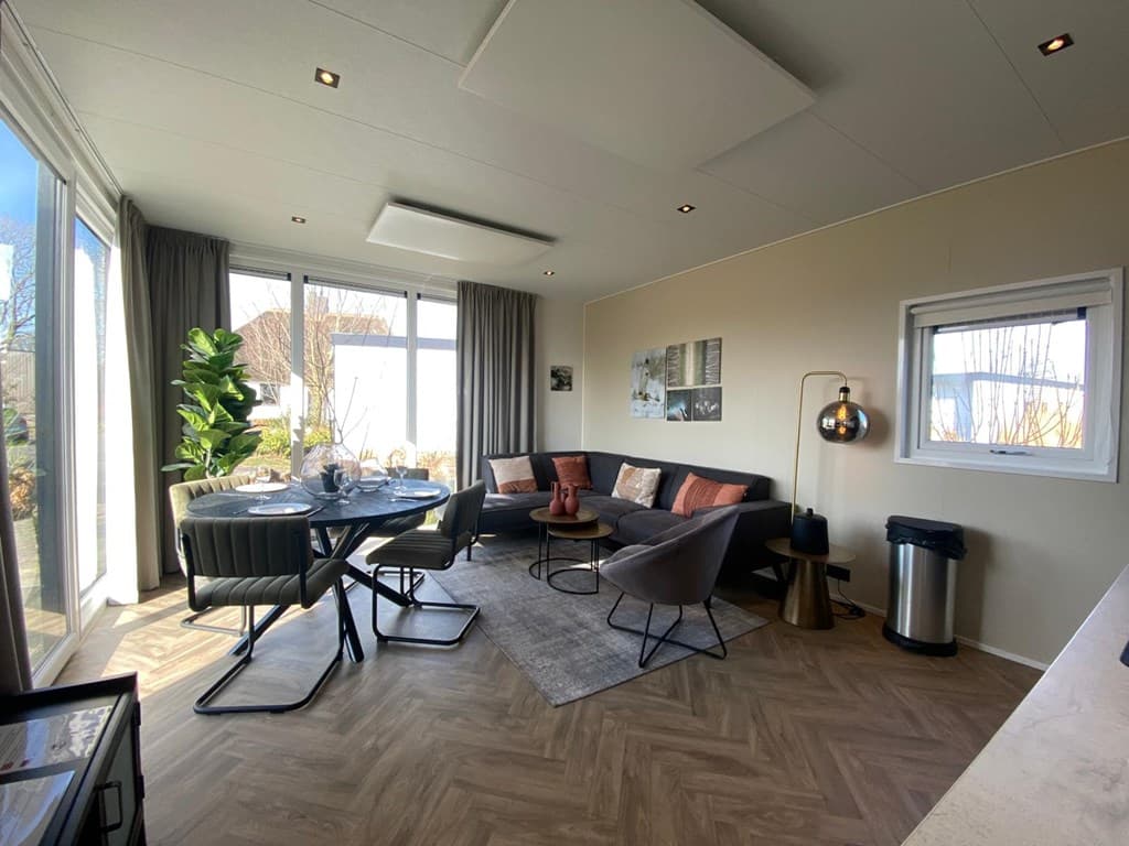 Moderne woonkamer met grote ramen, een eettafel, bank en fauteuils, lichte kleuren en een plantenbak in de hoek