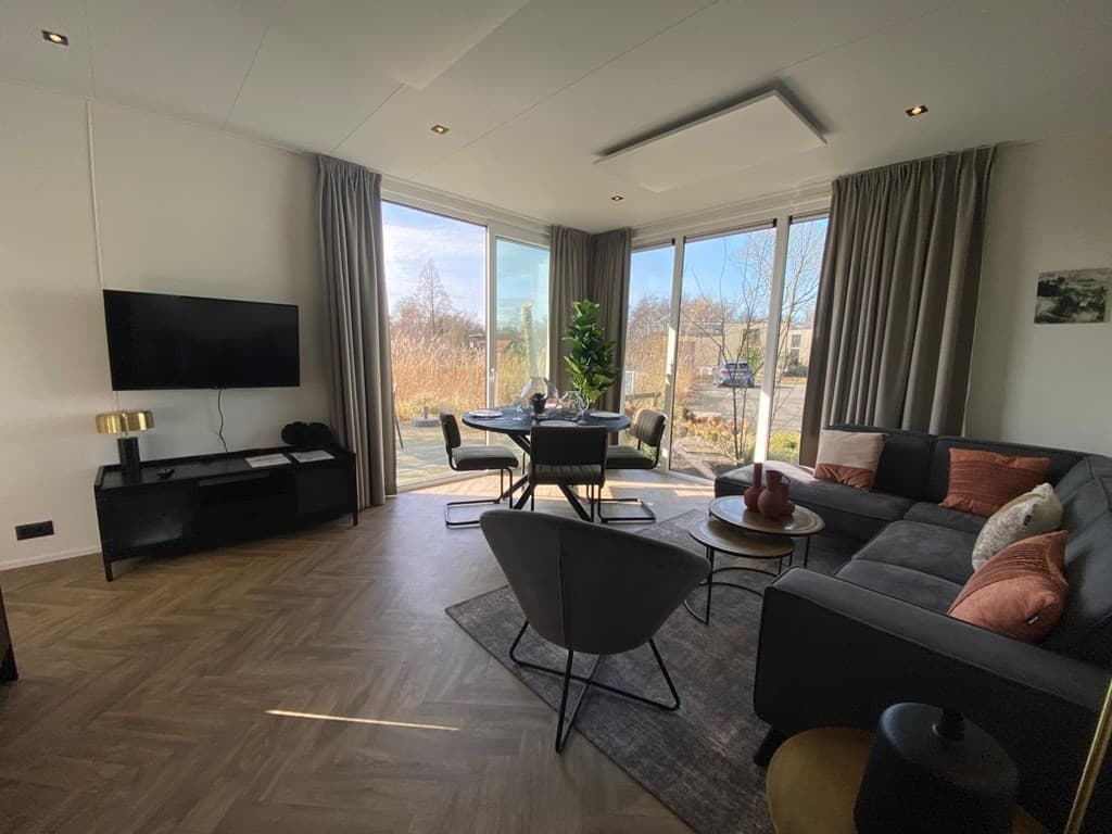 Moderne woonkamer met grote ramen, zithoek, eettafel en uitzicht op groen landschap in Vakantiepark De Wiedense Meren.
