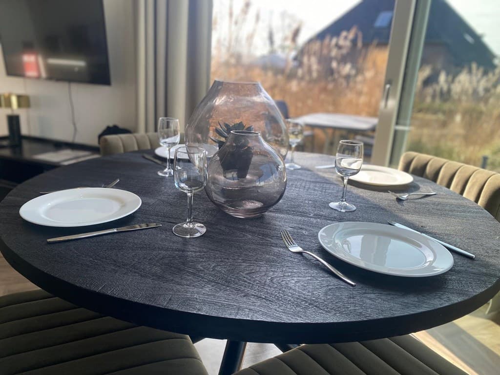 Eetkamertafel met glazen vaas en servies, omringd door stoelen, met uitzicht op een tuin en op de achtergrond een vakantiehui