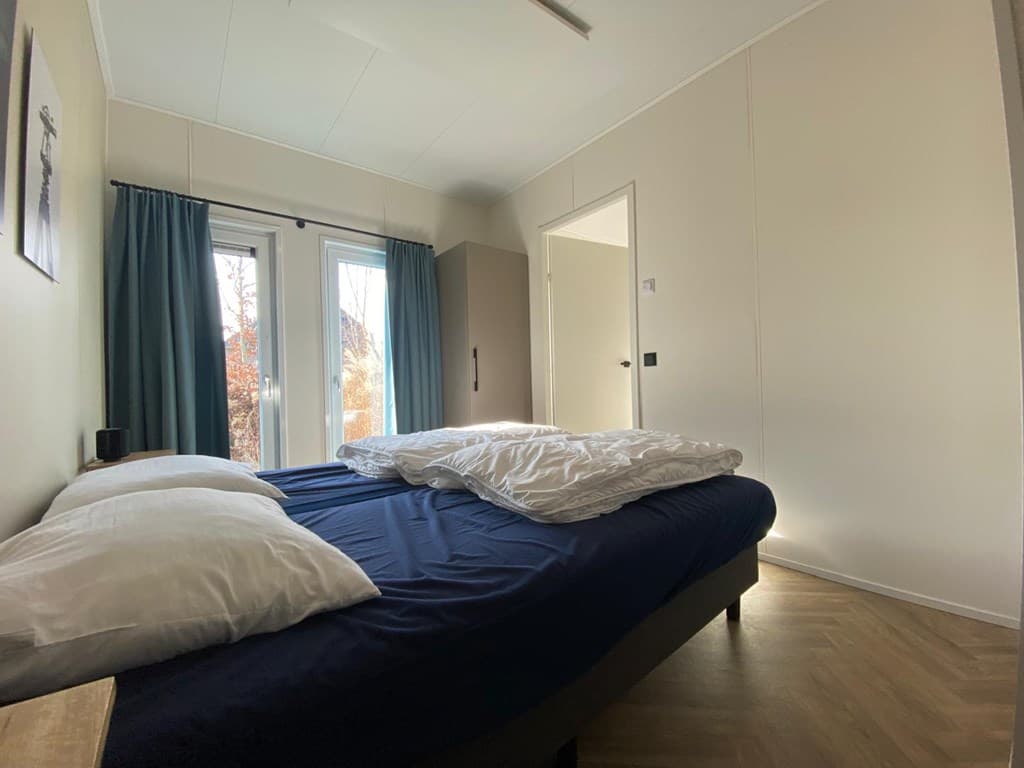 Slaapkamer met tweepersoonsbed, ramen met gordijnen en toegang naar een gang in de achtergrond