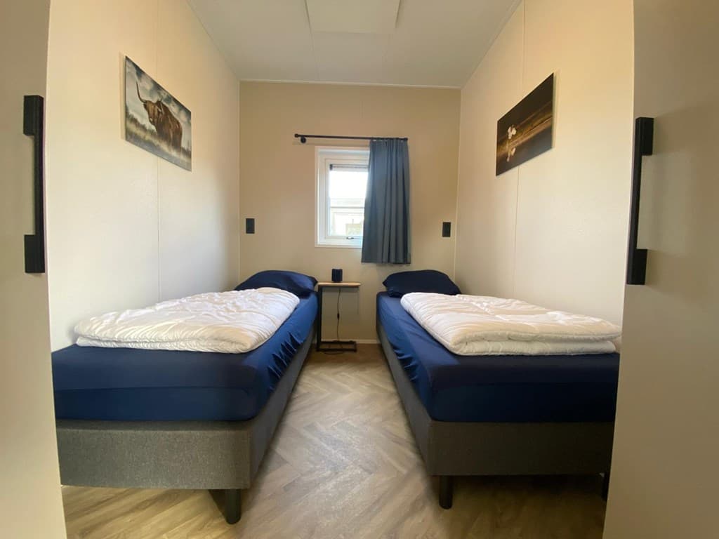 Slaapkamer met twee eenpersoonsbedden, raam met gordijn en kunst aan de wanden. Houten vloer en lampje zichtbaar.