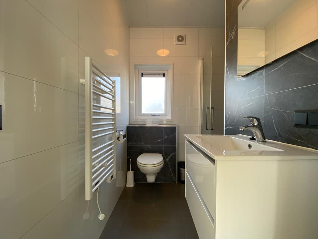 Moderne badkamer met wastafel, toilet en grote spiegel, witte en donkere betegeling met radiator aan de muur.