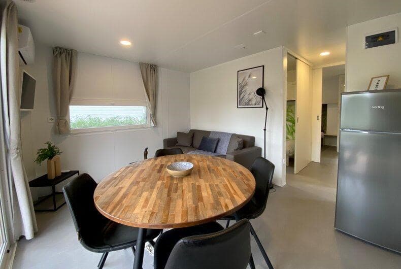In een lichte woning met een ronde houten eettafel, stoelen, bank en een wandtelevisie.