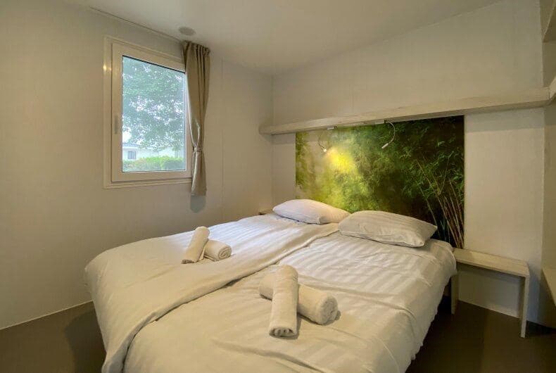 Slaapkamer met tweepersoonsbed, lichte wand, en grote raam met uitzicht op groen. Handdoeken op het bed gelegd.