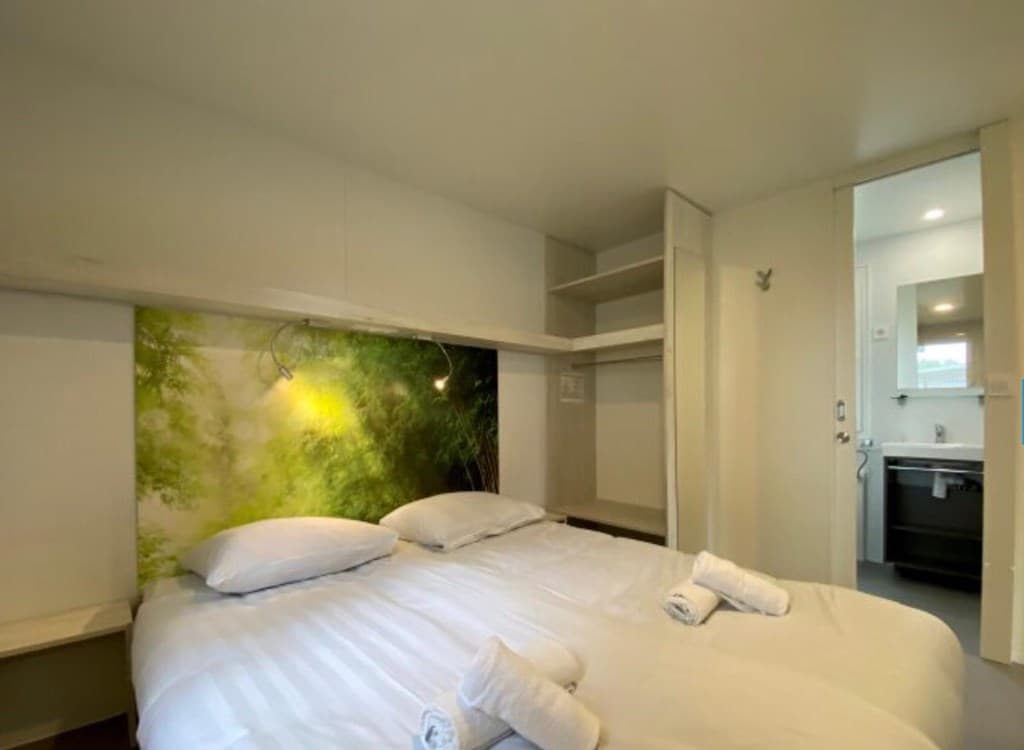 Slaapkamer met twee aparte bedden, kast, en een muurdecoratie met groen boslandschap. Keukenruimte zichtbaar in de achtergron