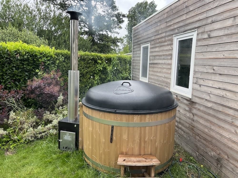 Houten hot tub met een dampende schoorsteen naast een moderne vakantiewoning en groene tuinbeplanting.