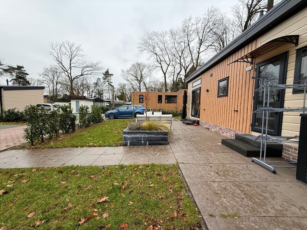 Buitenzicht op een bungalow met houten gevel, groene grasvelden en een parkeerplaats, omringd door bomen en andere woningen