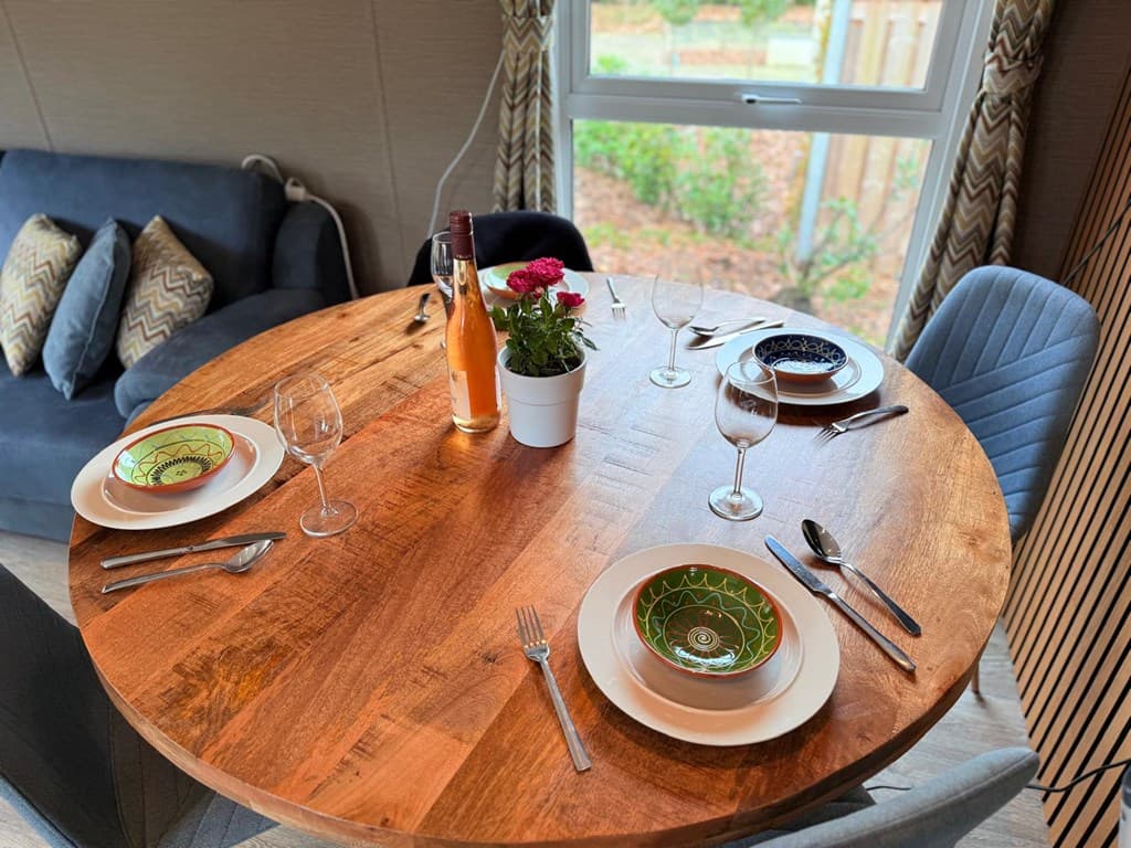 Eettafel met houten blad, gedekte borden, glazen, een fles en een bloemenpotje bij een raam met uitzicht op groen.