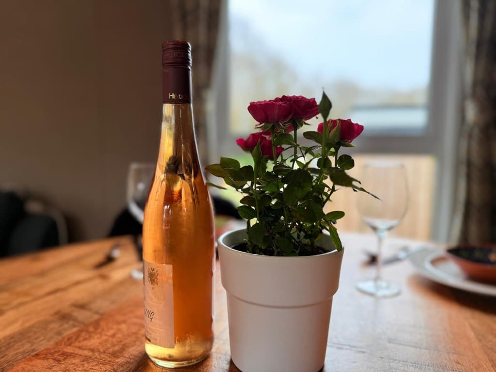 Glazen tafelschikking met een fles rosé en een witte pot met rozen, uitzicht op het water op de achtergrond.
