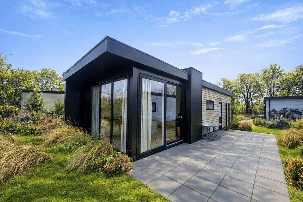 Moderne vakantiewoning met grote glazen muren en een betegeld terras, omgeven door groen en bloemen.