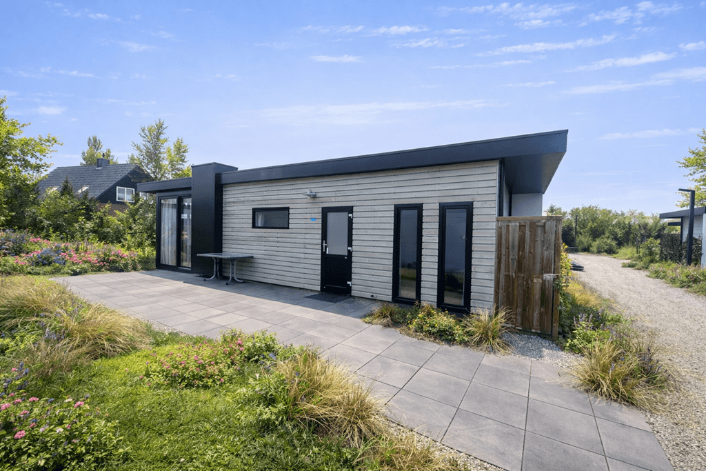 Moderne vakantiewoning met houten gevel en zwarte accenten, omgeven door bloemen en gras, met een betegeld pad.