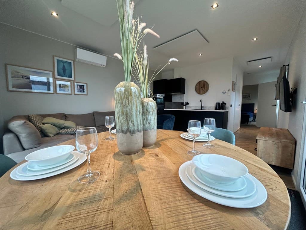 Eettafel met servies en glazen, uitzicht op een moderne woonkamer met een bank en een open keuken.