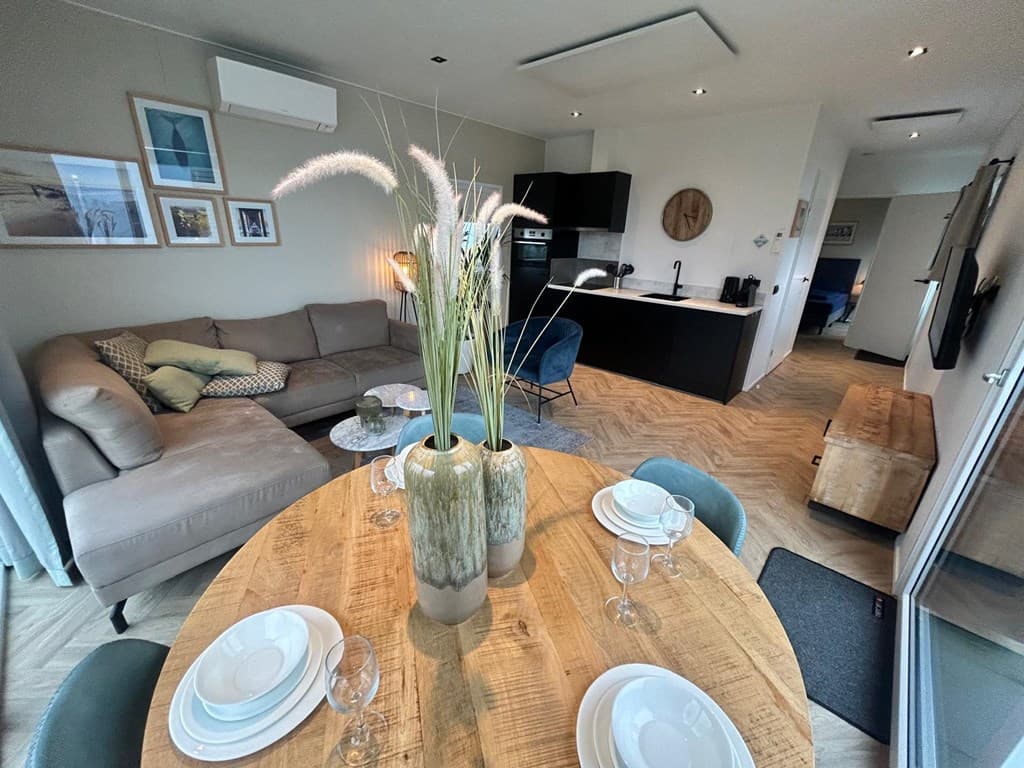Moderne woonkamer met open keuken, ronde eettafel, bank en decoratieve planten in een vakantiehuis.