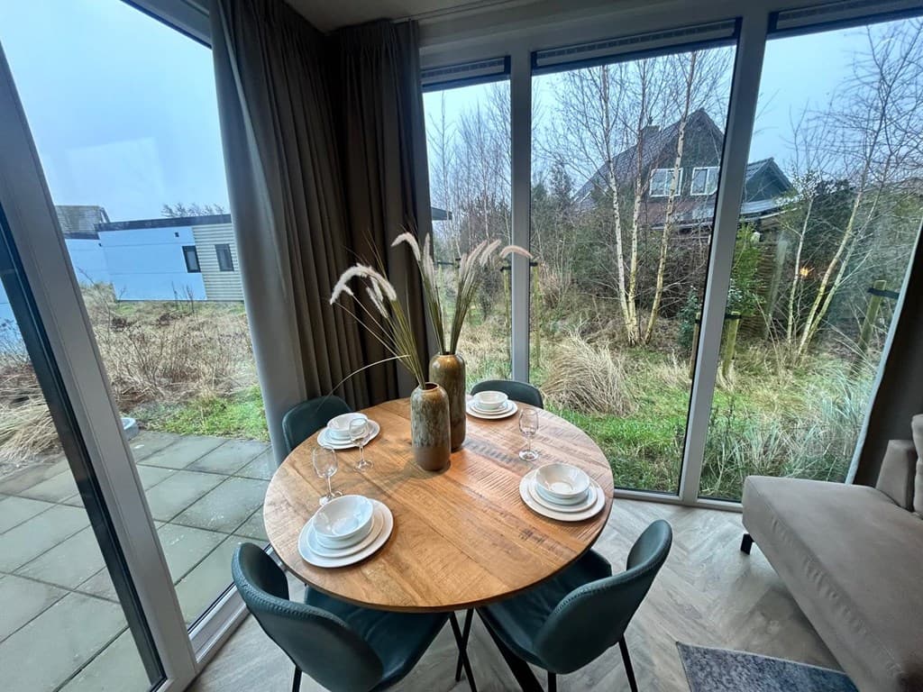 Eettafel met vier stoelen en decoratieve vaas, uitzicht op een groene omgeving met hoge grasplanten en huizen in de achtergro