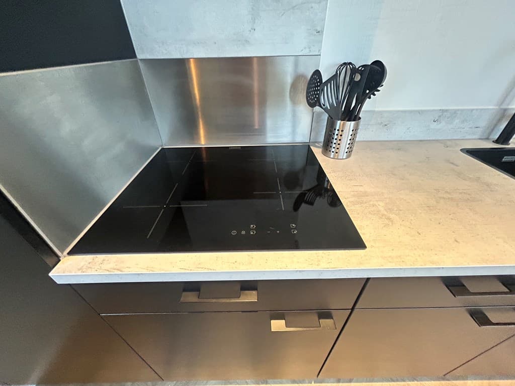 Inductiekookplaat met roestvrijstalen achterwand en keukengerei in de container op een stenen werkblad.