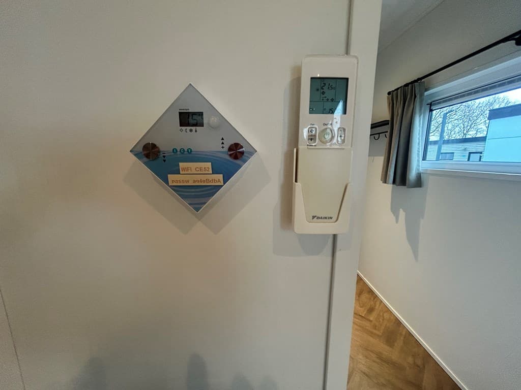 Thermostaat en bedieningspaneel aan de wand bij een raam met gordijnen en een houten vloer zichtbaar.