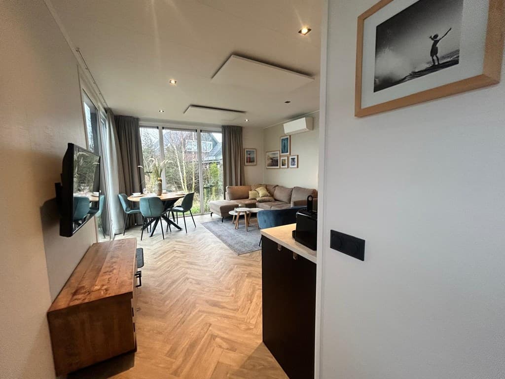 Moderne woonkamer met zithoek, eettafel en grote ramen die uitkijken op de tuin. Houten vloer en lichte muren.