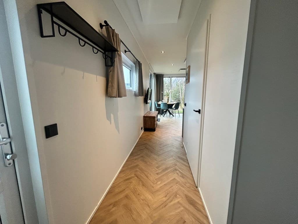 Hal met houten vloeren, wandhaakjes en zicht op een licht ingerichte woonkamer met eethoek en ramen naar buiten.