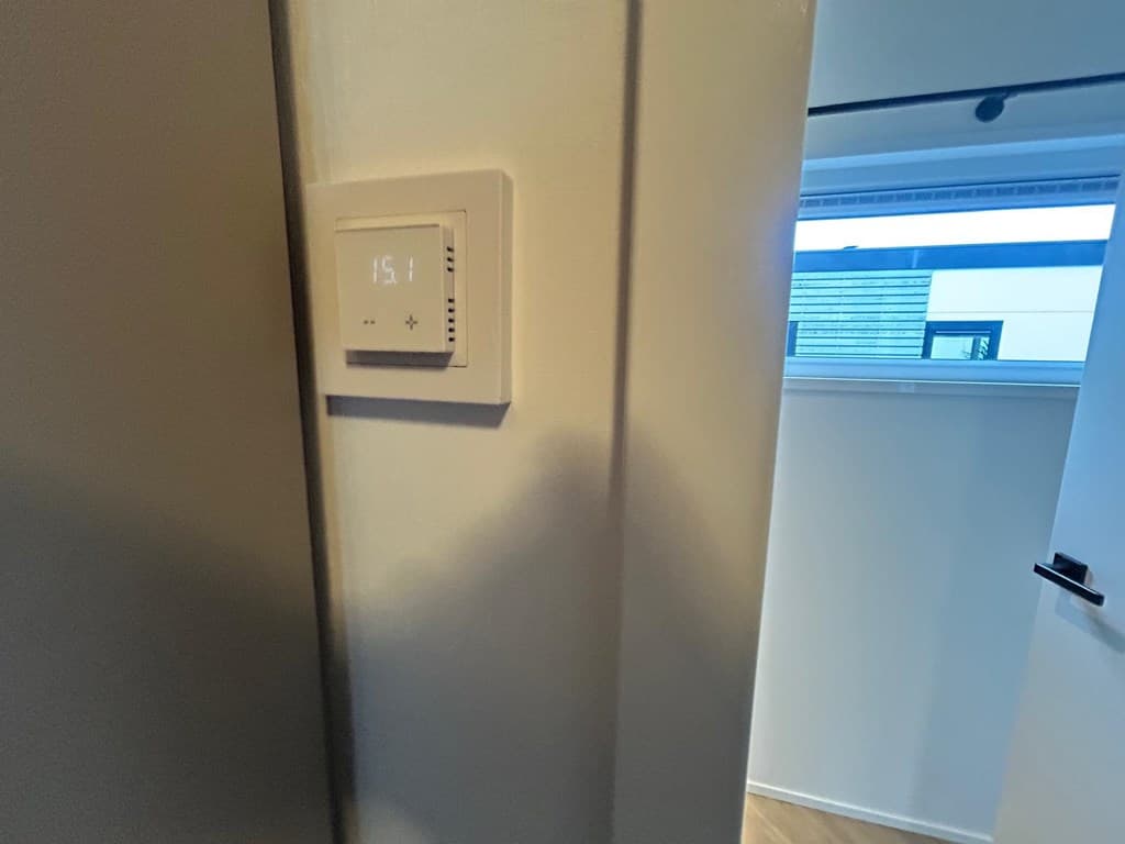 Thermostaat aan de muur bij een deur, zicht op een raam met uitzicht op de buitenruimte.