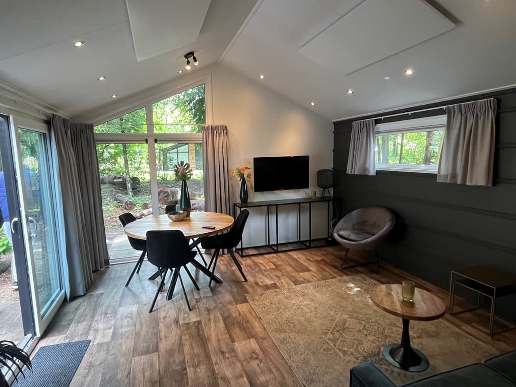 Gezellige woonkamer met eettafel, stoelen, zithoek, televisie en veel natuurlijk licht vanuit de ramen.