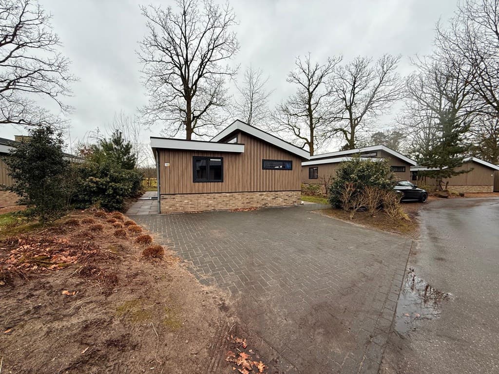 Vakantiehuis met houten gevel en grote ramen, gelegen tussen bomen op een grindpad met parkeerplek voor de deur