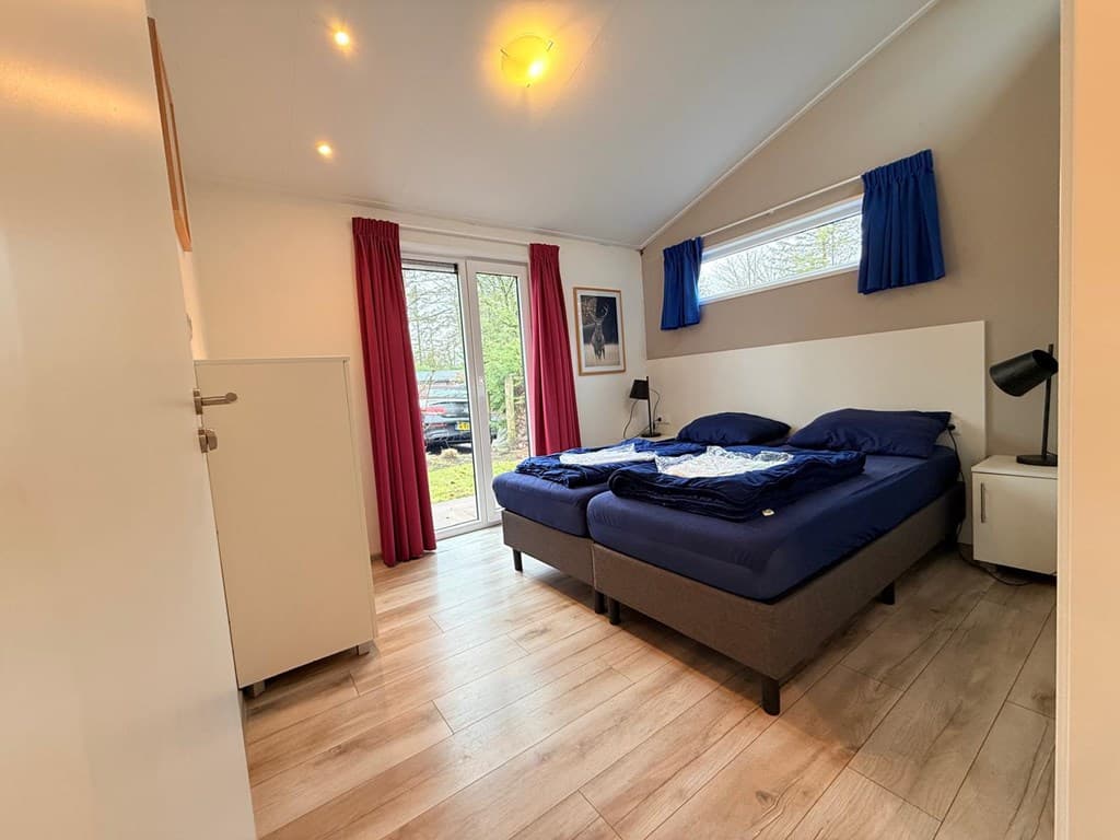 Slaapkamer met twee aparte bedden, grote ramen met blauwe en rode gordijnen, en een moderne inrichting