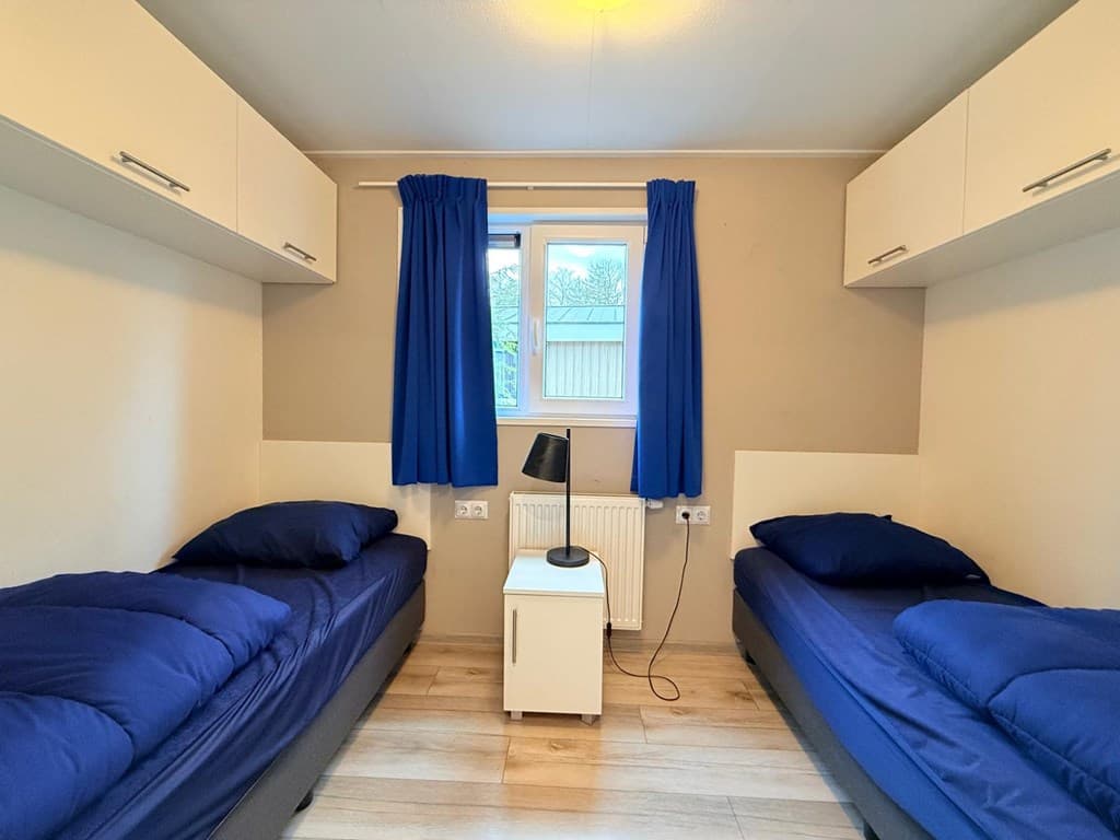 Slaapkamer met twee eenpersoonsbedden, kastjes en een raam met blauwe gordijnen.