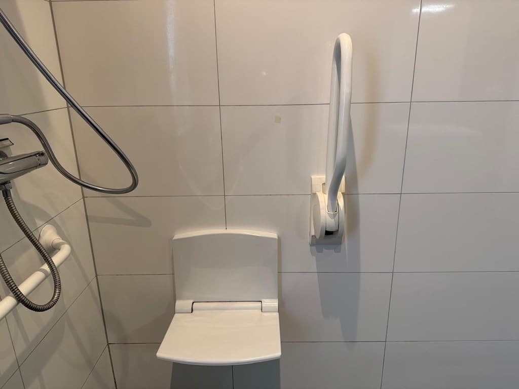 Witte wand met tegelwerk en een douchestoel aan de muur met een steunbeugel.