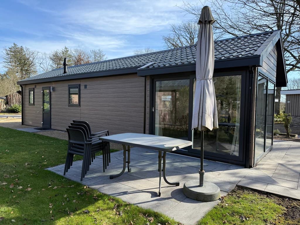 Vakantiehuis met een terras, een tafel met stoelen en een parasol, omgeven door groen en bomen.