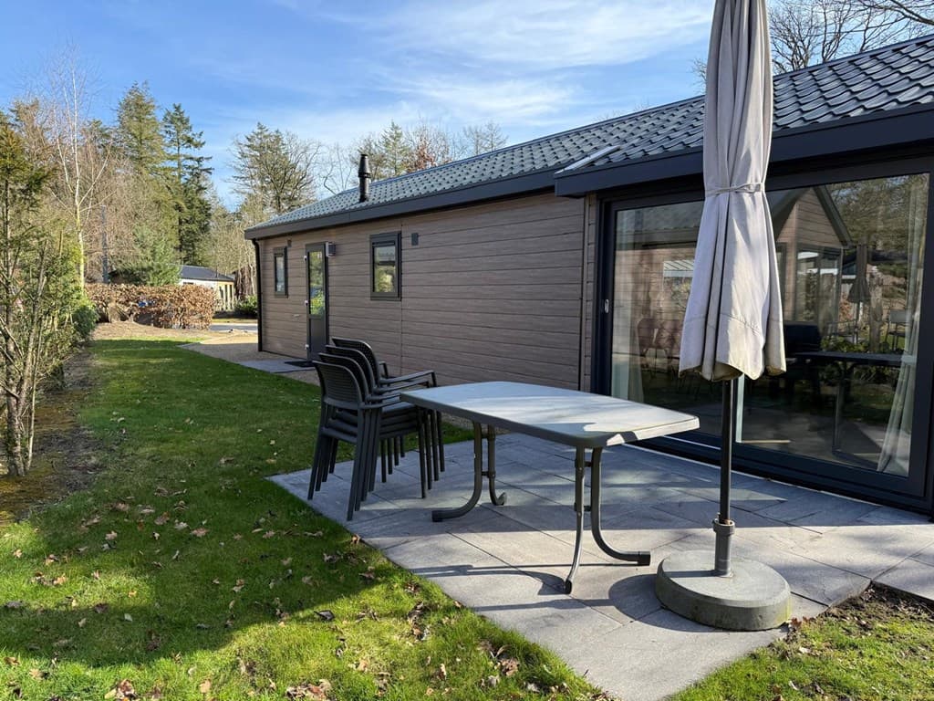 Terras met een tafel, stoelen en parasol bij een vakantiewoning omgeven door gras en bomen