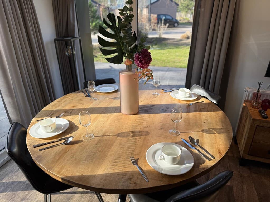 Ronde houten eettafel met servies, glazen en een vaas met bloemen in een lichte ruimte met grote ramen.