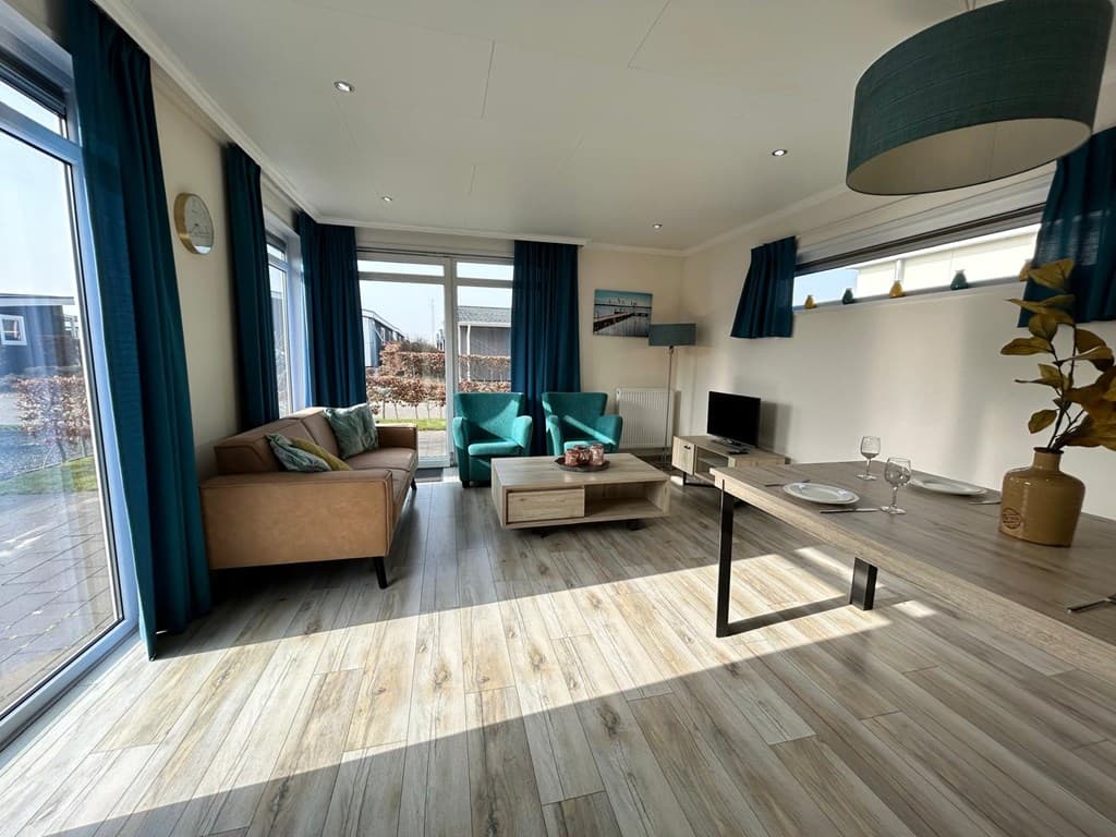 Moderne woonkamer met lichtinval, bank, twee stoelen, tv en eettafel met vaat en decoratie.