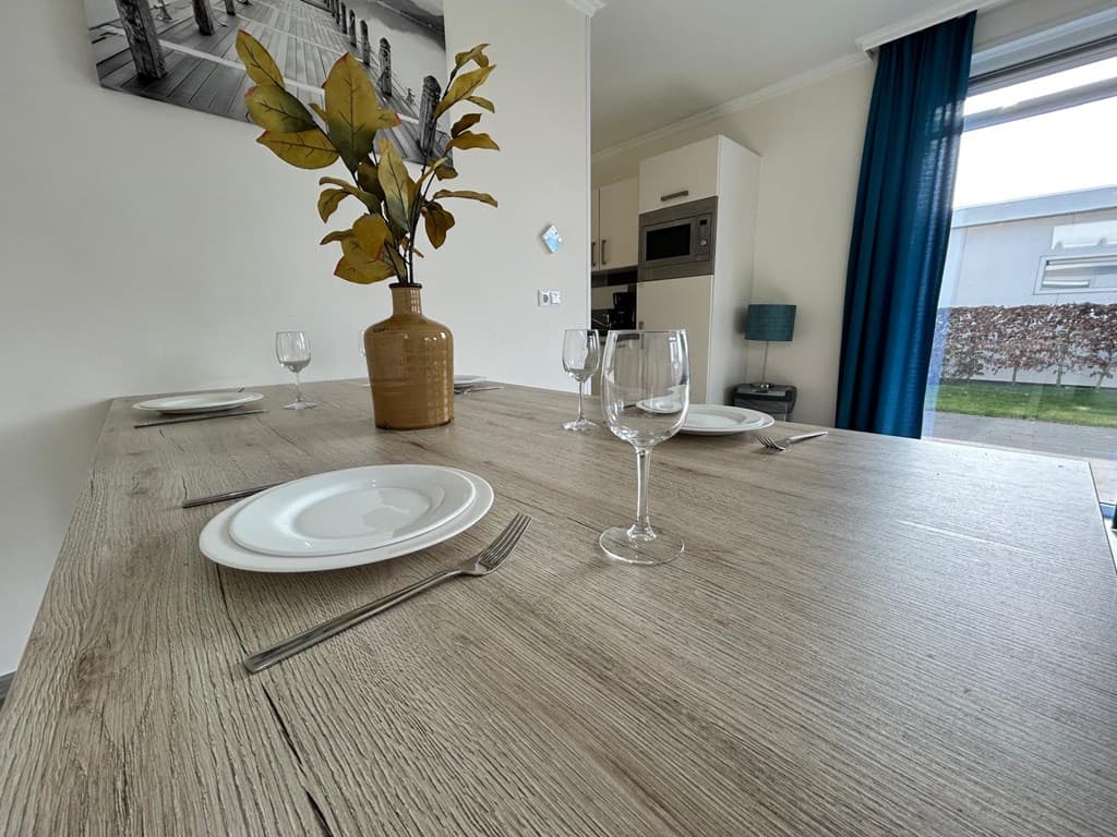 Eettafel met vier borden, glazen en een vaas met bladeren in een lichte ruimte met keuken op de achtergrond