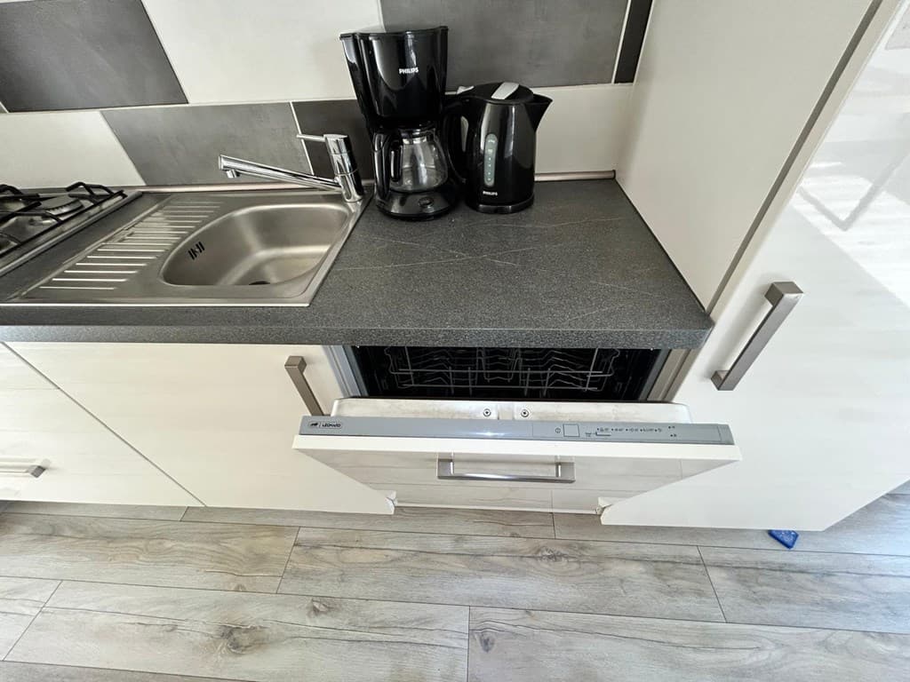 Keuken met aanrecht, opvangbak, koffiezetapparaat, waterkoker en open vaatwasser in lichtgekleurde kasten.