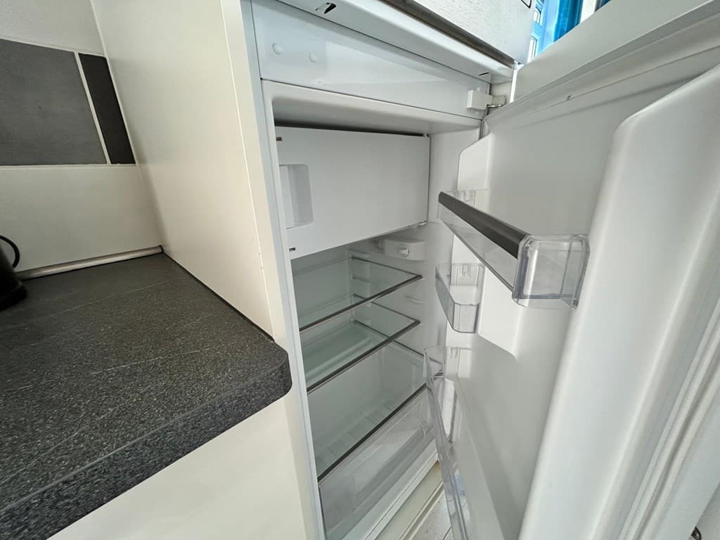Open koelkast met verschillende glazen planken en een transparante lade, witte binnenkant en een zwart aanrechtblad ernaast